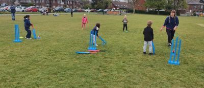 Small children and an instructor on grass playing cricket with soft bats and wickets