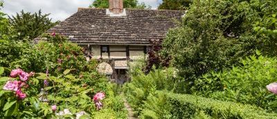 The Priest House stands in a traditional cottage garden. Greenery with pink flowers in front of the house.